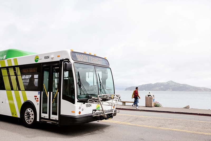 A modern public transit bus numbered 1504 stops at a waterfront location with mountains visible across the bay, while a pedestrian waits nearby on the promenade.