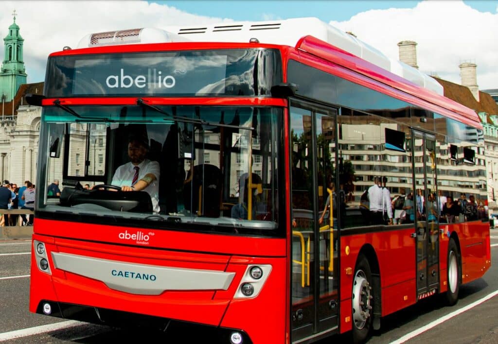 Red Abellio London double-decker bus with Caetano branding operating in an urban city center, featuring a driver and passengers visible through windows.
