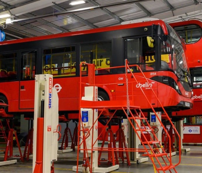 Red Abello bus number 8154 undergoes maintenance in a depot with red service platforms and lifting equipment visible alongside the vehicle.