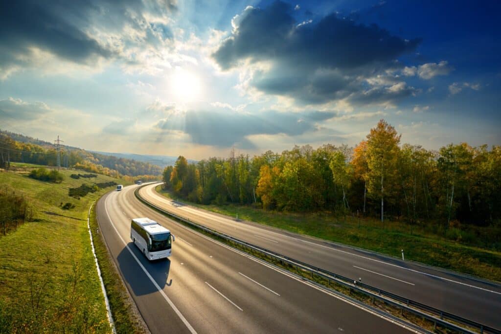 A white bus travels on a curved highway through rolling forested countryside during autumn, with dramatic sunlit clouds overhead and green hillsides flanking the road.
