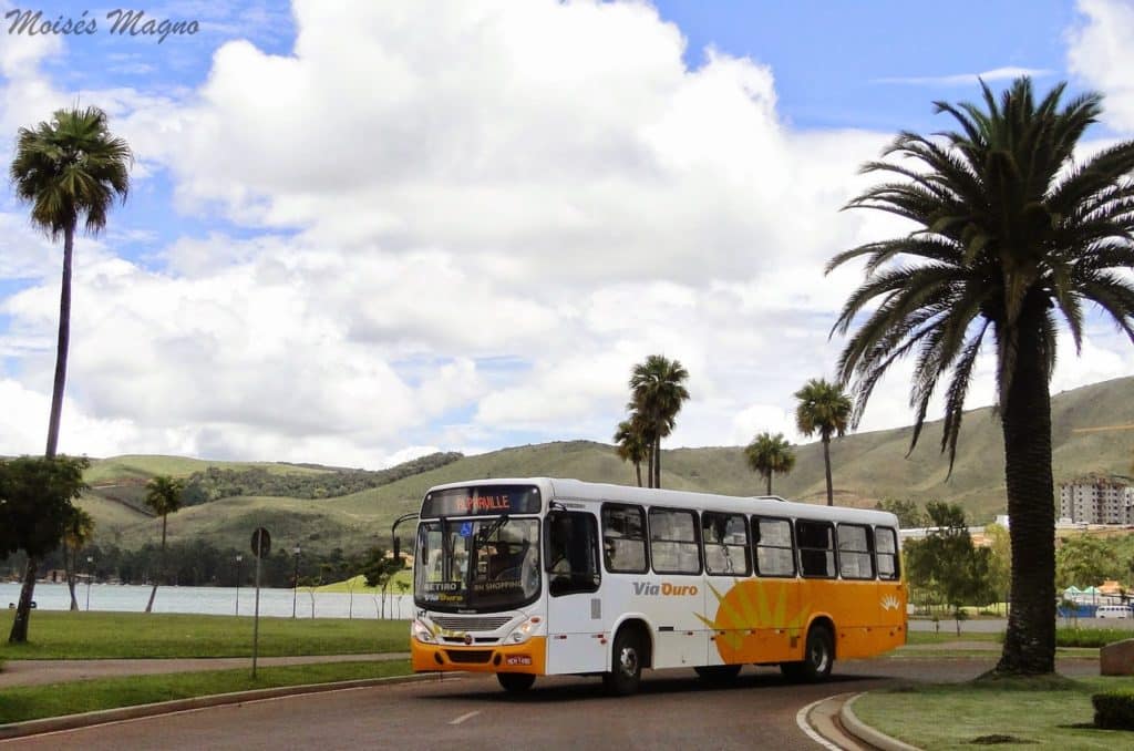 Via Ouro bus in orange and white livery parked beside a lake with palm trees and green hills in the background, Brazil.