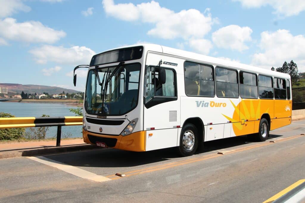 Via Ouro white and yellow bus parked by a scenic lakeside overlook with mountains and blue sky in the background. Brazilian passenger transportation service.