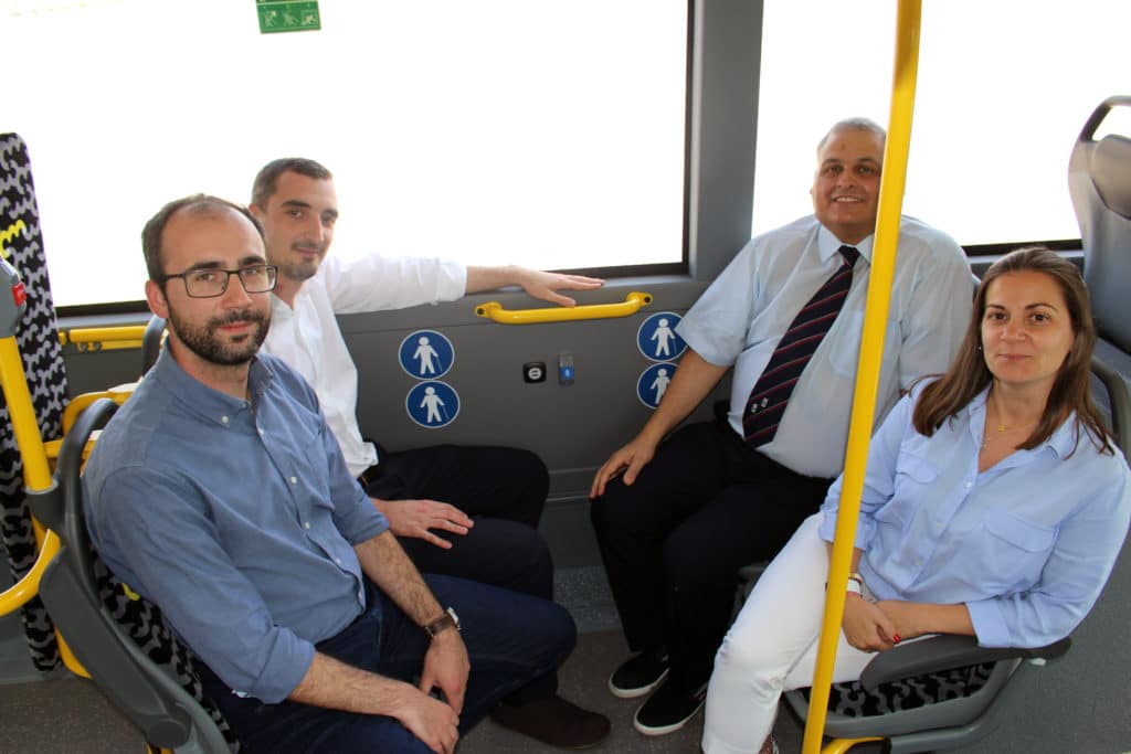 Five professionals sit inside a modern public transit bus, smiling at the camera near the door area marked with pedestrian icons and yellow safety handles.