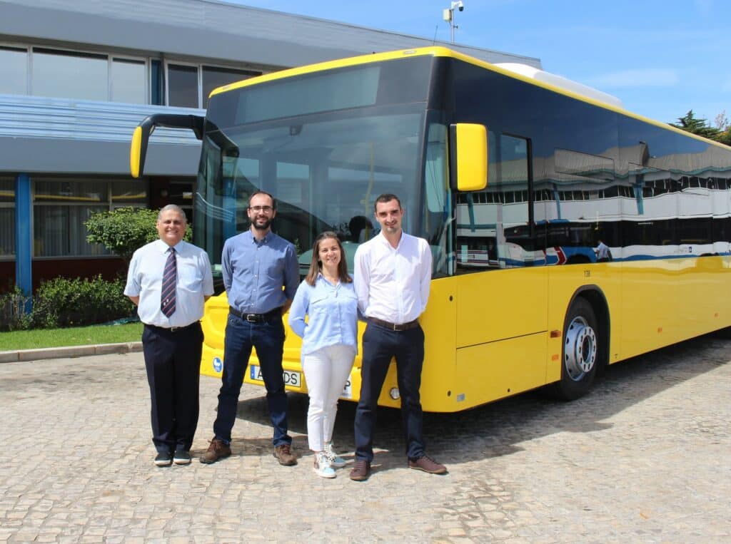 Four professionals stand in front of a yellow public transit bus at a transportation facility. The team poses confidently beside the modern vehicle on a sunny day.
