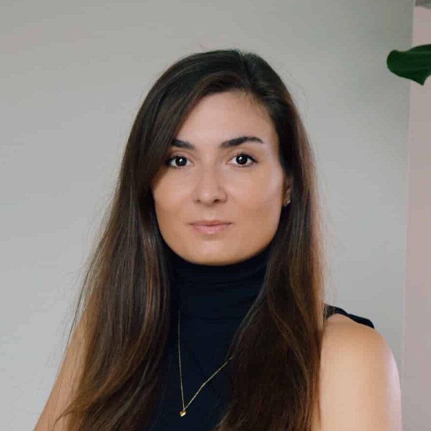 Professional portrait of a woman with long dark hair and defined eyebrows, wearing a black turtleneck and gold pendant necklace against a neutral background.