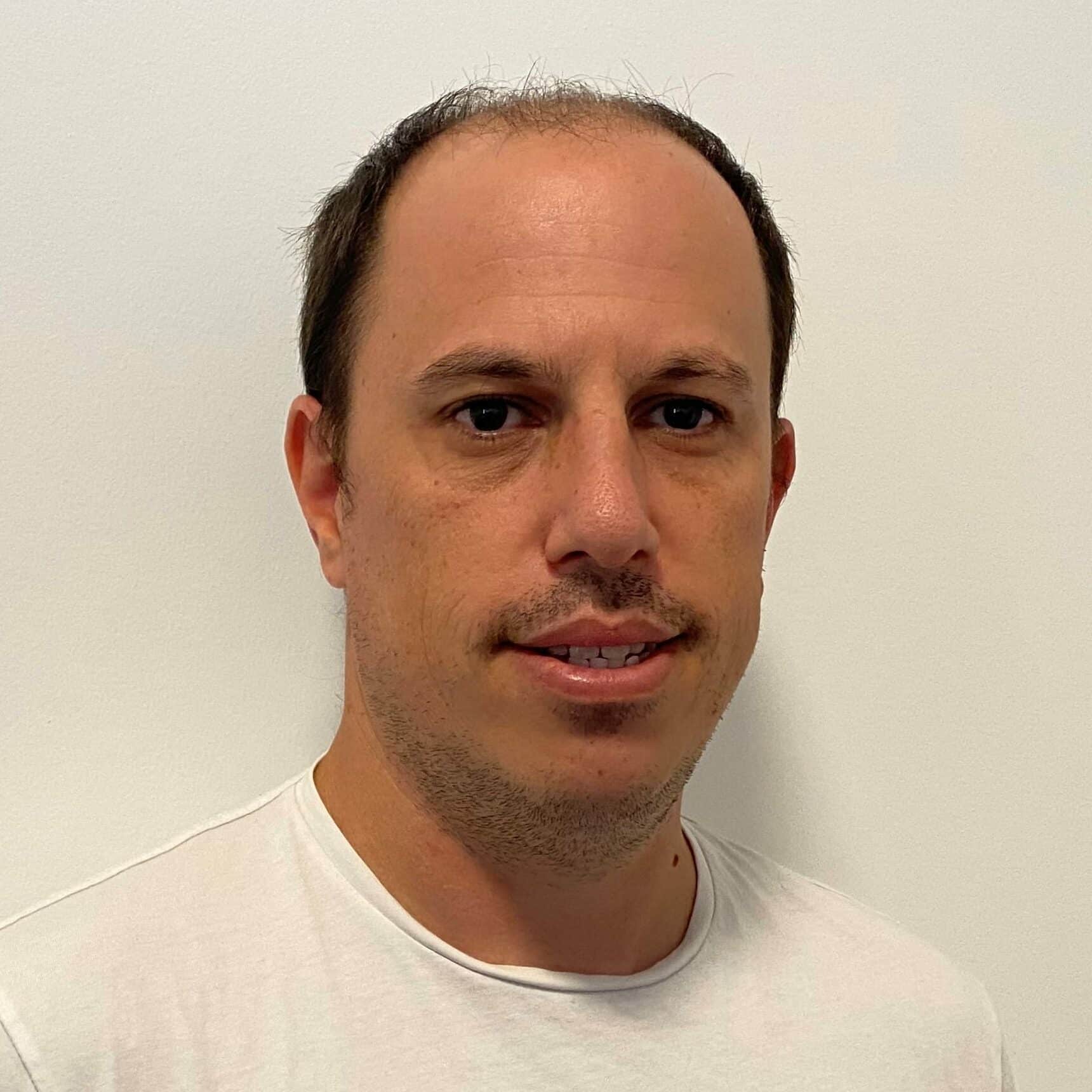 Portrait of a middle-aged man with receding dark hair and facial stubble wearing a white t-shirt against a neutral background.