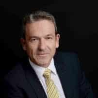 Professional headshot of a middle-aged man wearing a dark suit and yellow striped tie against a black background, displaying a composed expression suitable for business or corporate use.