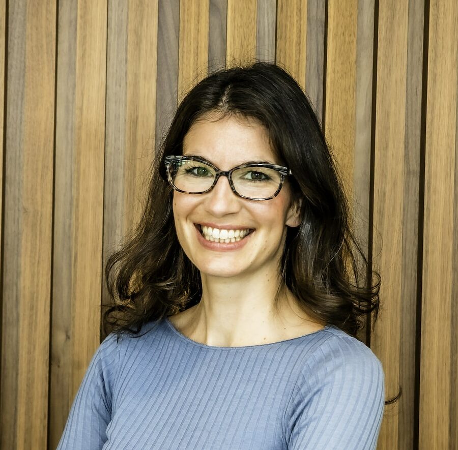 Portrait of a smiling woman wearing glasses and a light blue ribbed shirt against a wooden vertical slat background.