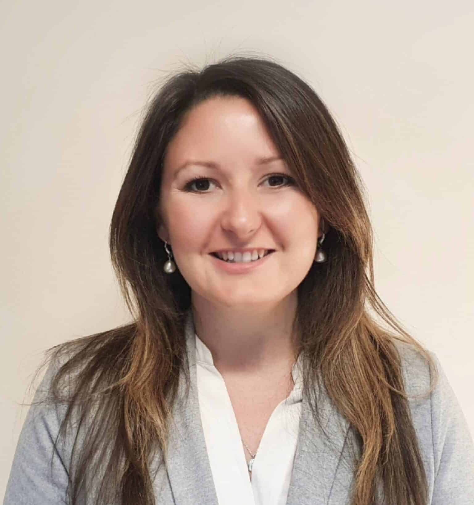 Professional headshot of a smiling woman with long brown hair wearing a gray blazer and white blouse against a neutral background.