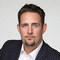 Professional headshot of a man wearing a dark pinstriped suit and white dress shirt against a neutral gray background, ideal for business or corporate use.