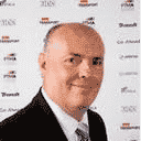 Professional headshot of an older man wearing a dark suit jacket and tie against a branded event backdrop with various logos visible.