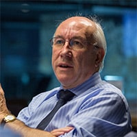 Professional man wearing glasses and blue shirt speaks during indoor presentation or meeting with blurred background.