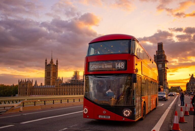 Red London double-decker bus heading to Clerkenwell Green crosses Westminster Bridge at sunset, with the Houses of Parliament and Big Ben illuminated in the background.