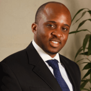 Michael Nsereko Professional portrait of a man in formal business attire wearing a black suit and navy tie, smiling at the camera against a neutral background with a potted plant visible.