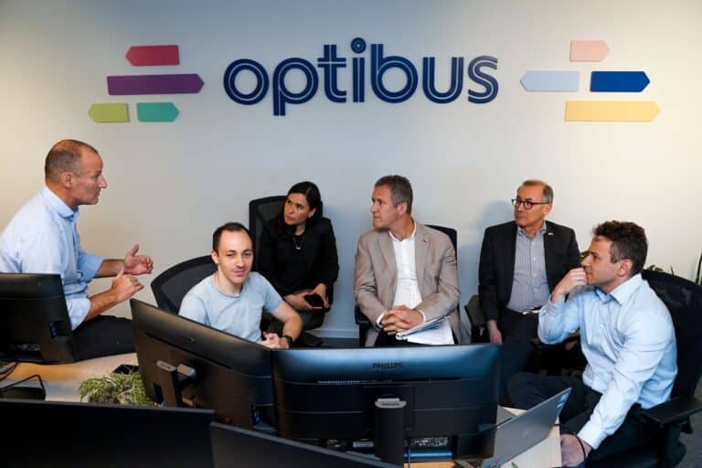 Team meeting at Optibus office with six professionals gathered around a computer monitor, discussing business strategy in a collaborative workspace.