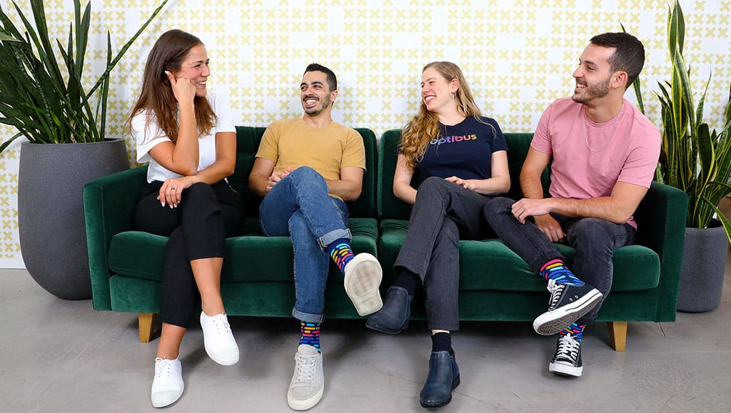 Four young adults sit together on a green velvet couch, smiling and chatting in a modern office setting with patterned wallpaper and potted plants.