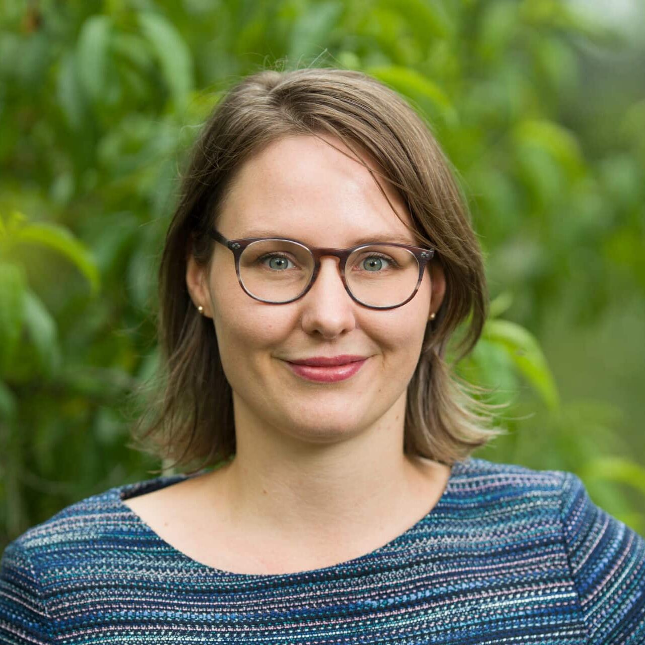 Woman wearing glasses and a blue striped sweater smiles at the camera against a blurred green foliage background in an outdoor portrait photograph.