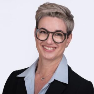 Professional headshot of a smiling woman wearing glasses and a black blazer over a light blue shirt, photographed against a white background.