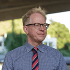 Professional man wearing glasses and a blue shirt with red striped tie, photographed outdoors with a blurred green background suggesting a patio or deck setting.