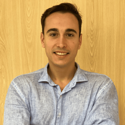 Professional headshot of a smiling man wearing a light blue button-up shirt against a wooden background. He appears friendly and approachable in this corporate portrait photograph.