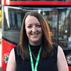 Smiling woman wearing a green lanyard badge stands in front of a red bus, appearing at an outdoor event or conference venue.
