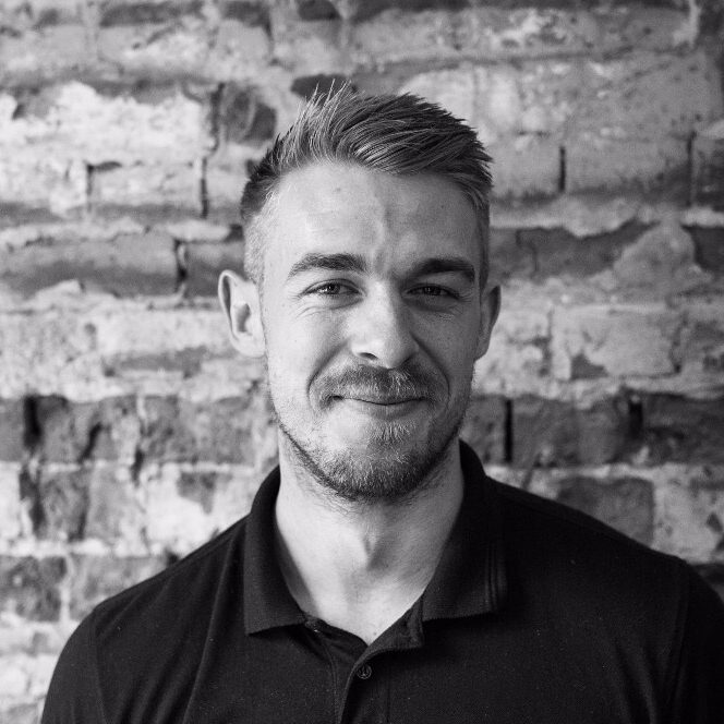 Professional headshot of a man with styled blonde hair and facial hair wearing a black polo shirt against a weathered brick wall background.