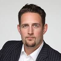 Professional headshot of a man wearing a dark pinstriped blazer and white dress shirt, photographed against a neutral gray background with confident expression and styled dark hair.
