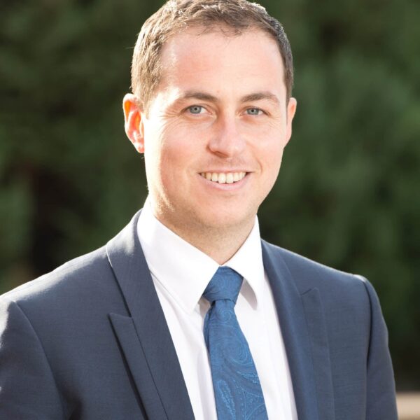 Professional headshot of a smiling man wearing a dark navy suit, white dress shirt, and blue patterned tie against a blurred green outdoor background.