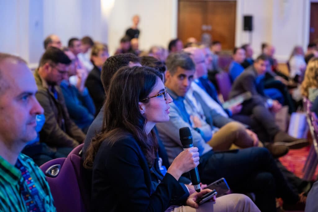 Woman in glasses speaks into microphone at community event, addressing seated audience in indoor venue with purple lighting.