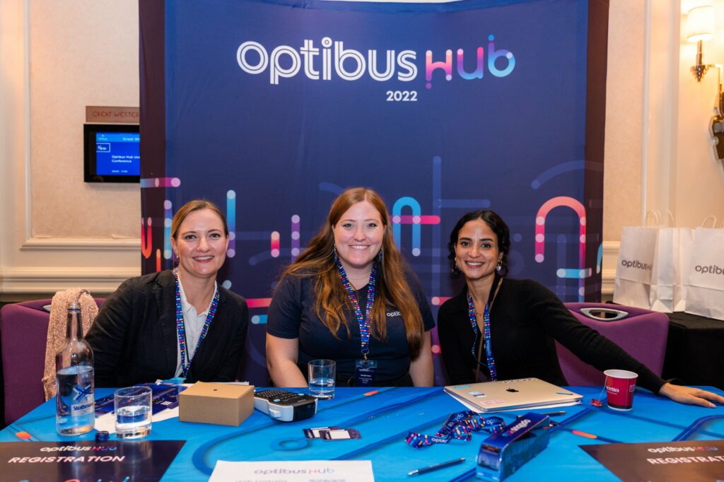 Three professional women wearing lanyards sit at a registration table at the Optibus Hub 2022 conference event, smiling at the camera.