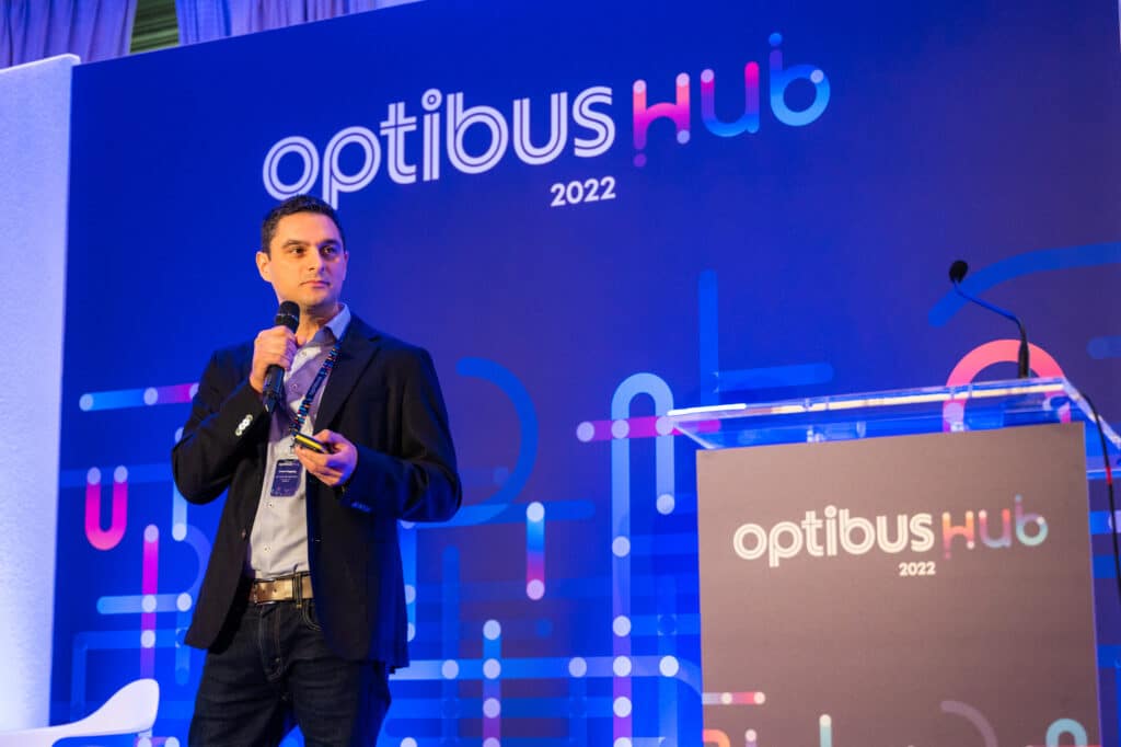 A professional speaker presents at the Optibus Hub 2022 conference, holding a microphone while standing before a vibrant blue branded backdrop with geometric designs and pink accent elements.