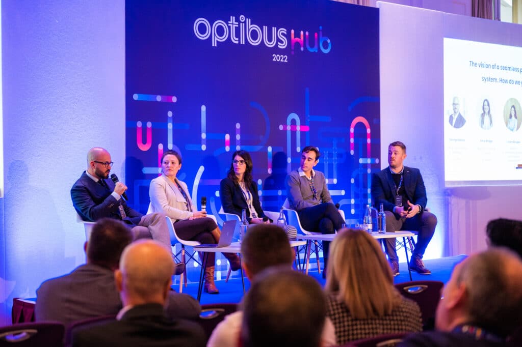 Panel discussion at OptiBus Hub 2022 conference features five professionals seated on stage discussing seamless payment systems, with audience members visible in foreground.