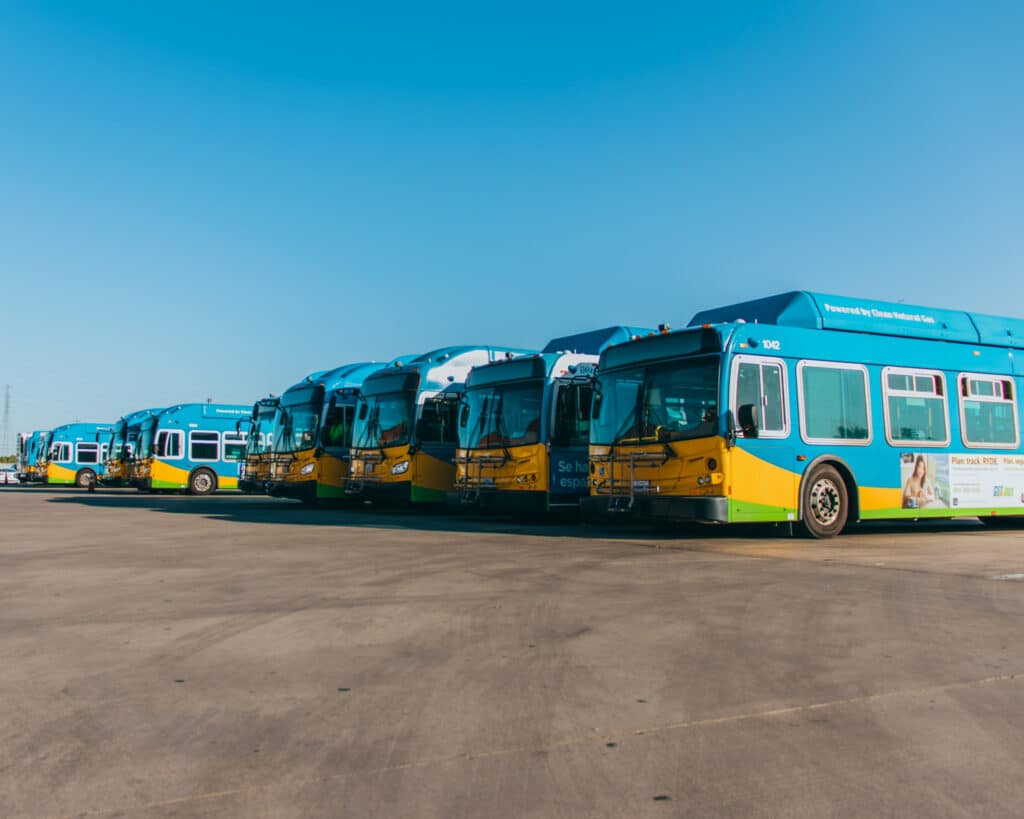 A fleet of blue, yellow, and green hybrid buses lined up in a row on an asphalt parking lot under clear skies, showcasing eco-friendly public transportation vehicles.