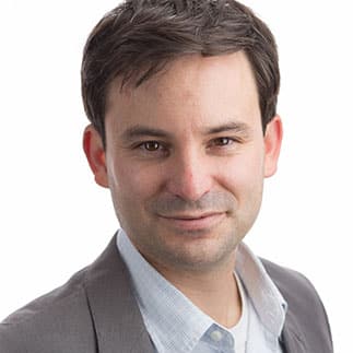Professional headshot of a middle-aged man wearing a gray blazer and light blue checkered shirt against a white background, suitable for business or corporate use.