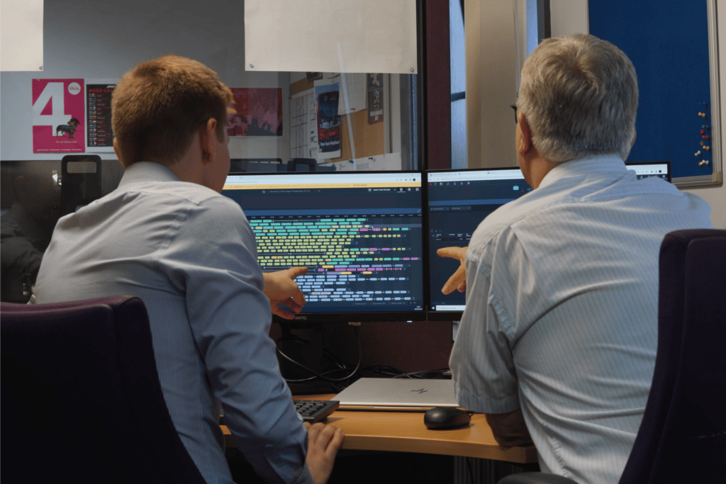 Two professionals review colorful data visualization on dual monitors in an office workspace. The younger man in a light blue shirt and older man in gray analyze what appears to be a complex