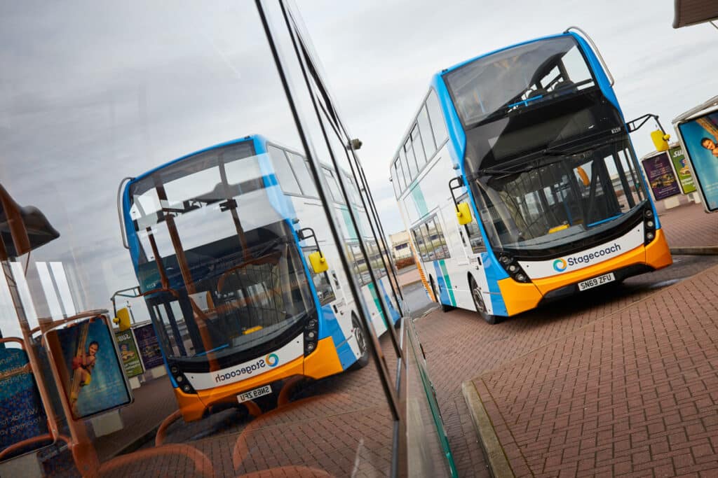 Two blue and orange Stagecoach double-decker buses parked on a brick plaza under cloudy skies. The vehicles feature the company's distinctive livery with white and turquoise upper sections.