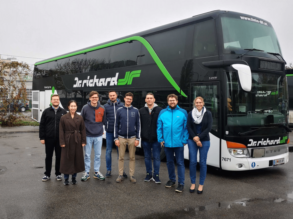 Eight people stand in front of a Dirichard coach bus with distinctive green and black livery, posing for a professional team photo in casual winter clothing.