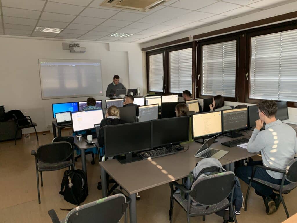 Computer science classroom with students seated at workstations with multiple monitors, focused on their screens while an instructor stands at the front near a projector display.