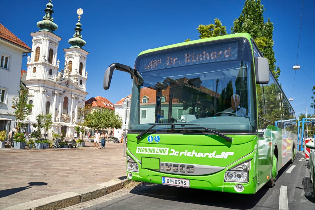 Bright green Verbund Linie bus parked in European town square with white baroque church featuring golden domes in background under clear blue sky.