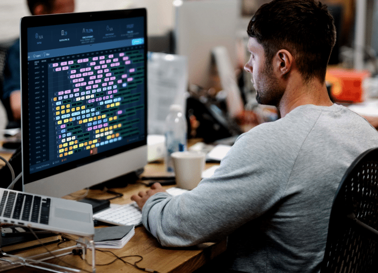 Developer analyzing colorful data visualization on computer monitor in a modern office workspace, wearing a gray shirt while reviewing complex code or analytics dashboard.