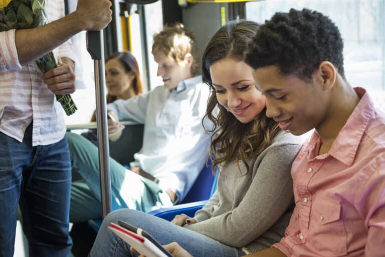 Young people riding public transportation, smiling while looking at a tablet together. A man holding flowers stands nearby as other passengers sit in the bus.