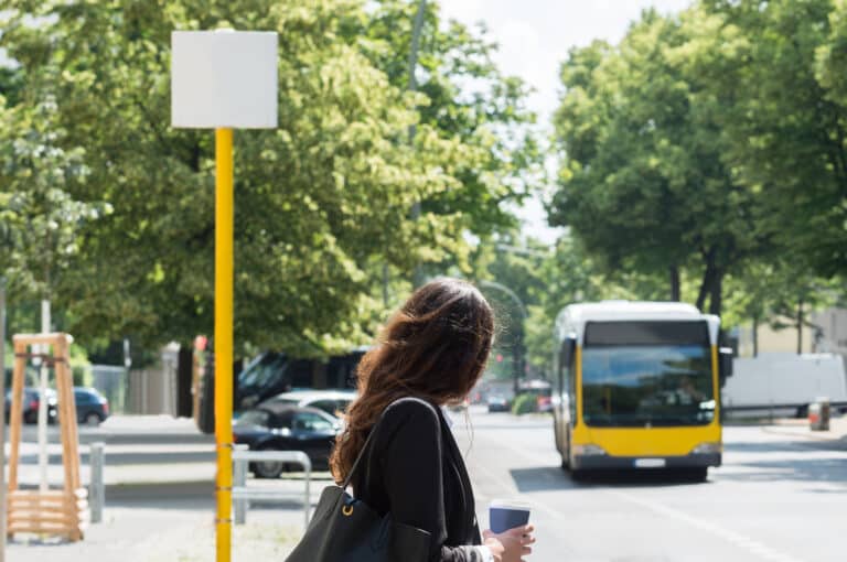 Woman waiting at a bus stop with a yellow pole and blank sign, holding a coffee cup as a yellow bus approaches on a tree-lined urban street.