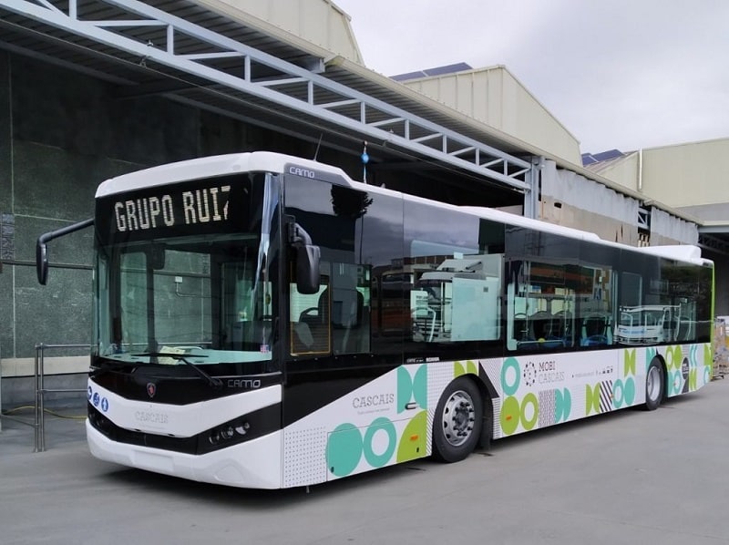 Modern black and white articulated bus operated by Grupo Ruit features turquoise and lime green accents on its lower panels, parked under industrial warehouse structures.