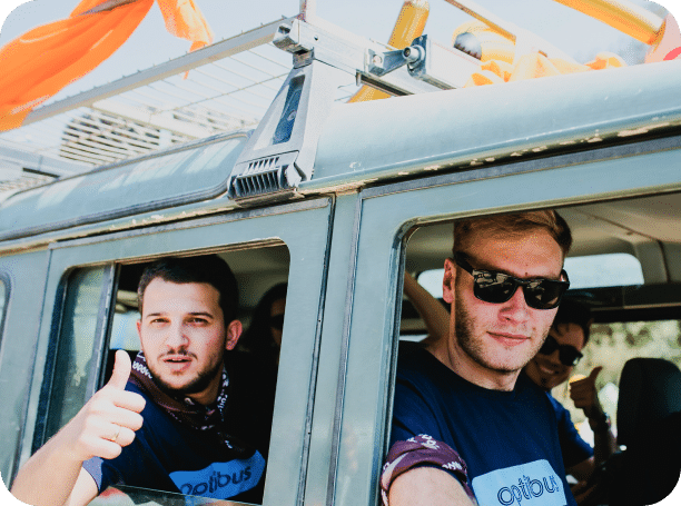 Two men in a helicopter give thumbs up and wear sunglasses during an aerial adventure tour, with orange parachutes visible on the aircraft roof.