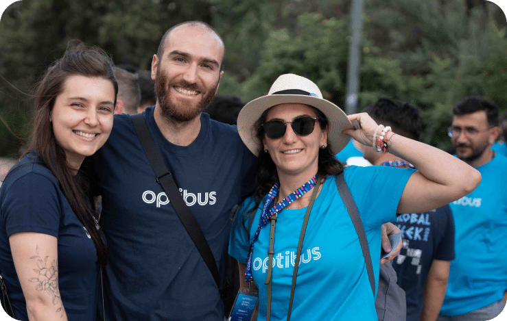 Three team members wearing Optibus branded shirts smile together outdoors at what appears to be a company event or gathering, with colleagues visible in the background.