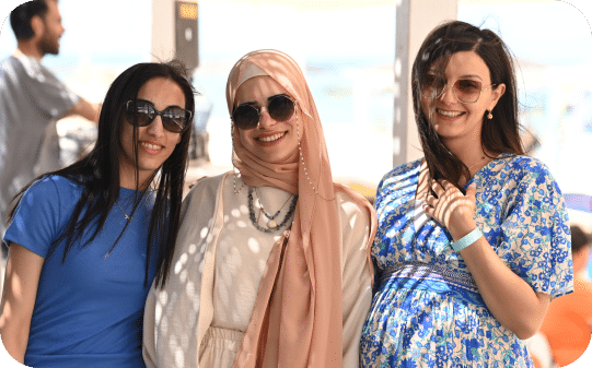 Three smiling women pose together indoors wearing sunglasses and casual summer clothing. They appear to be enjoying time at a social gathering or event.