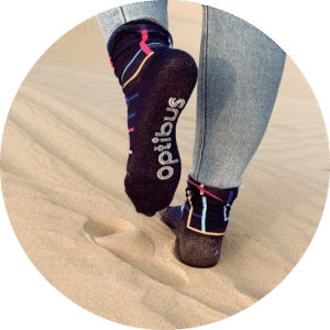 A person wearing dark socks with colorful stripes walks barefoot on sandy beach, displaying comfortable Opiöo brand compression socks designed for active wear.