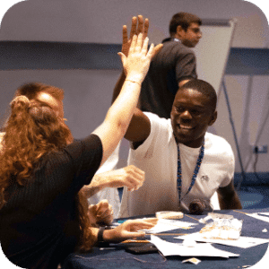 Professionals celebrate success with a high-five at a conference table during a networking event. A woman in black and a smiling man wearing a credential badge share an enthusiastic gesture of