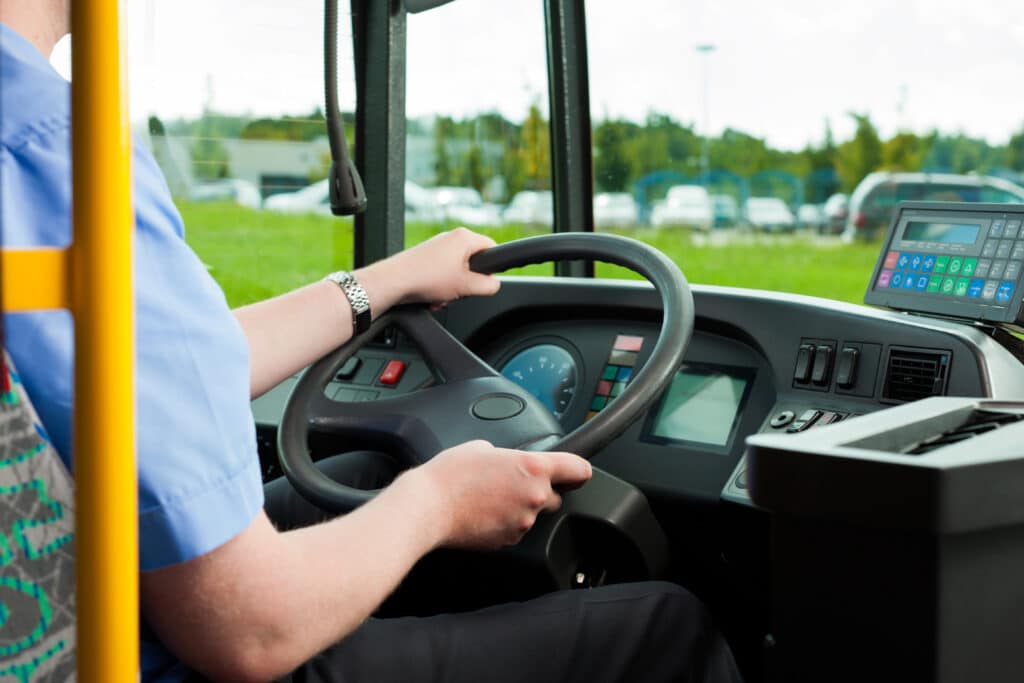 Bus driver gripping the steering wheel inside a transit vehicle, wearing a blue uniform and bracelet, with control panels and countryside visible through the windshield.
