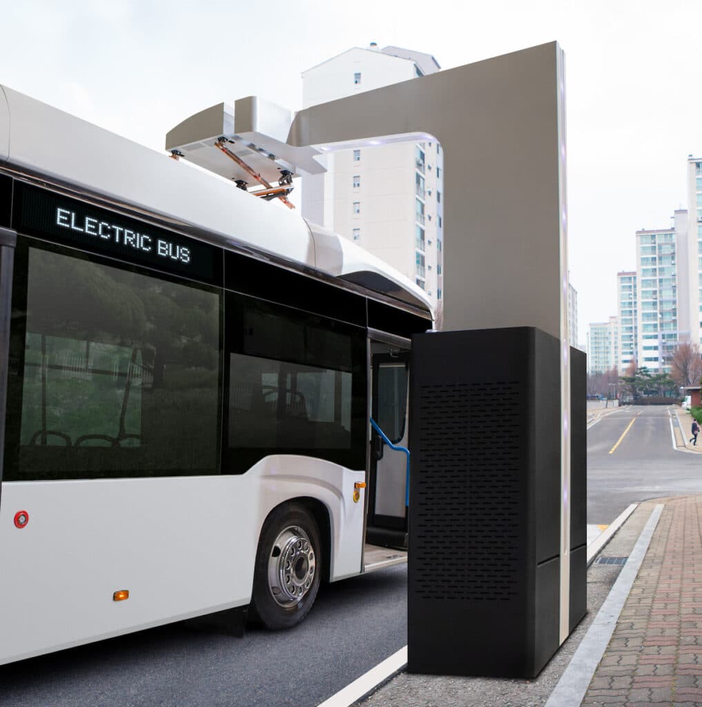 Electric bus charging at a overhead pantograph charging station in an urban setting with residential buildings in the background.